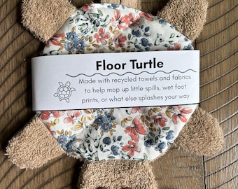 May include: A turtle-shaped cleaning cloth made of tan terry cloth with a floral patterned fabric center. It features two brown buttons as eyes and a paper band labeled Floor Turtle with text about its use for mopping up spills and wet footprints.