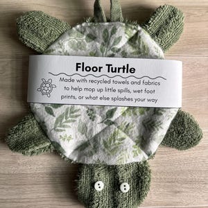 May include: A turtle-shaped cleaning cloth made of green terry cloth with a white and green leaf-patterned center. It features two white button eyes and a paper band labeled Floor Turtle with text about its use for spills and wet footprints.