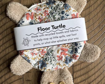 May include: A turtle-shaped cleaning cloth made of beige terry cloth with a floral patterned top. It features two yellow buttons as eyes and a hanging loop. A white paper band reads: Floor Turtle, Made with recycled towels and fabrics to help mop up little spills, wet foot prints, or what else splashes your way.