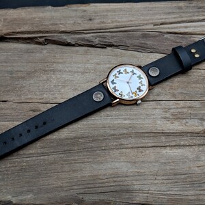 K&ouml;nnte beinhalten: Ein schwarzes Lederarmband mit einem ros&eacute;goldenen Zifferblatt, auf dem ein Kreis aus bunten Schmetterlingen zu sehen ist.
