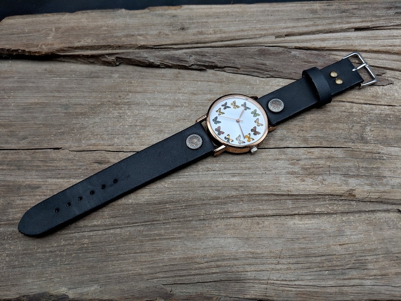 K&ouml;nnte beinhalten: Ein schwarzes Lederarmband mit einem ros&eacute;goldenen Zifferblatt, auf dem ein Kreis aus bunten Schmetterlingen zu sehen ist.