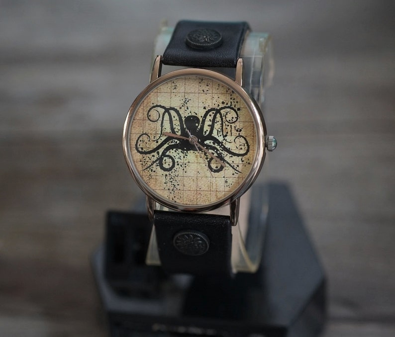 Pu&ograve; includere: Un orologio da polso in pelle nera con un quadrante rotondo di colore crema che presenta un'illustrazione di un polpo nero. Il quadrante dell'orologio ha un motivo a griglia e lancette nere.