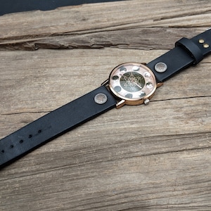 Pode incluir: Um rel&oacute;gio de pulso de couro preto com um mostrador redondo de ouro rosa com um design de fase lunar. O rel&oacute;gio est&aacute; sobre uma superf&iacute;cie de madeira.