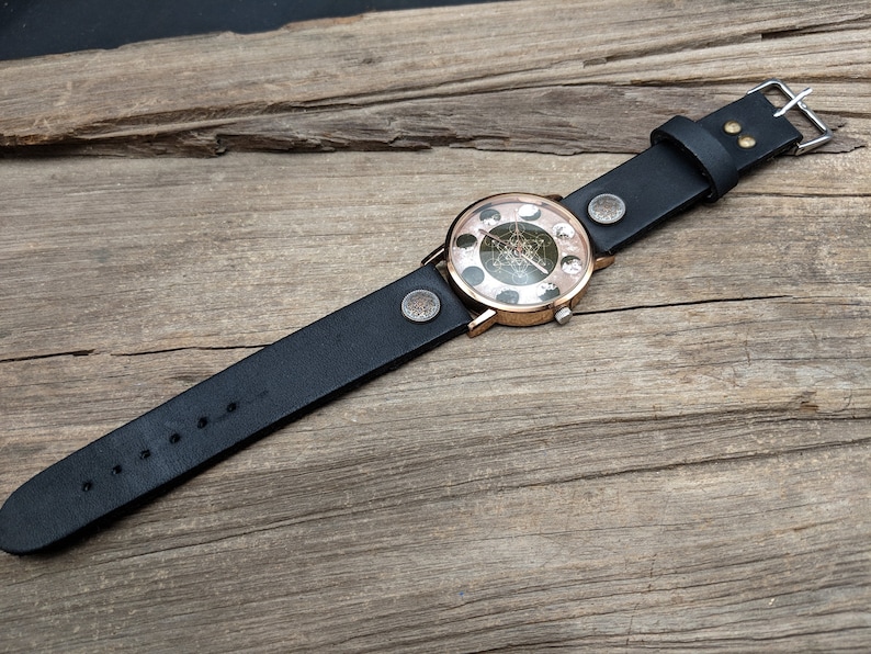 Pode incluir: Um rel&oacute;gio de pulso de couro preto com um mostrador redondo de ouro rosa com um design de fase lunar. O rel&oacute;gio est&aacute; sobre uma superf&iacute;cie de madeira.