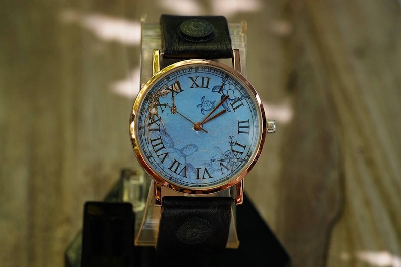 May include: A gold-tone wristwatch with a blue face and Roman numeral markings. The watch face features a seahorse design and a black leather band.