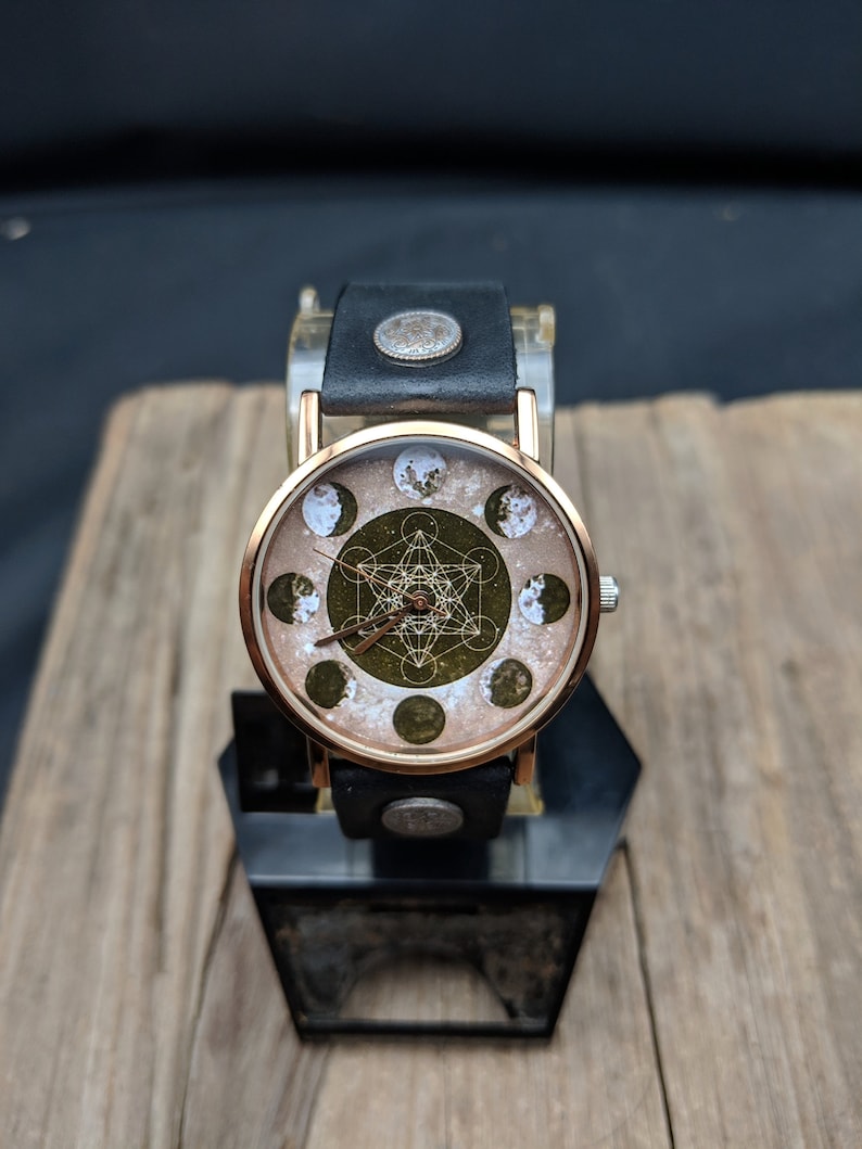 Pode incluir: Um rel&oacute;gio de pulso de ouro rosa com uma pulseira de couro preto. O mostrador do rel&oacute;gio tem um fundo branco com um design geom&eacute;trico verde e uma s&eacute;rie de fases lunares.