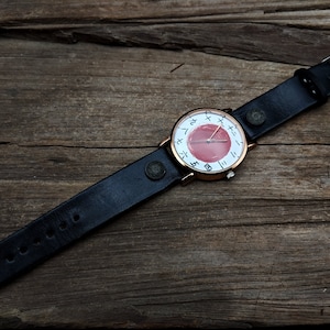May include: A wristwatch with a black leather band and a round face. The watch face features a red center with white markings and a gold-colored bezel. The watch is set against a rustic wooden background.