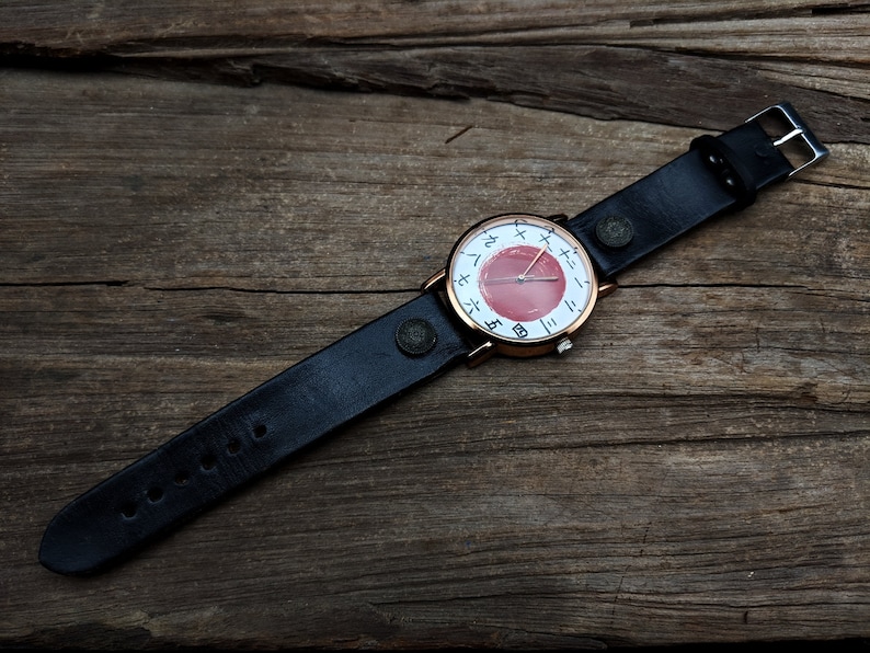 May include: A wristwatch with a black leather band and a round face. The watch face features a red center with white markings and a gold-colored bezel. The watch is set against a rustic wooden background.