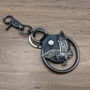 May include: A silver key chain with a leather eagle charm. The eagle is in flight and has a detailed design. The key chain has a silver clip and a silver ring.