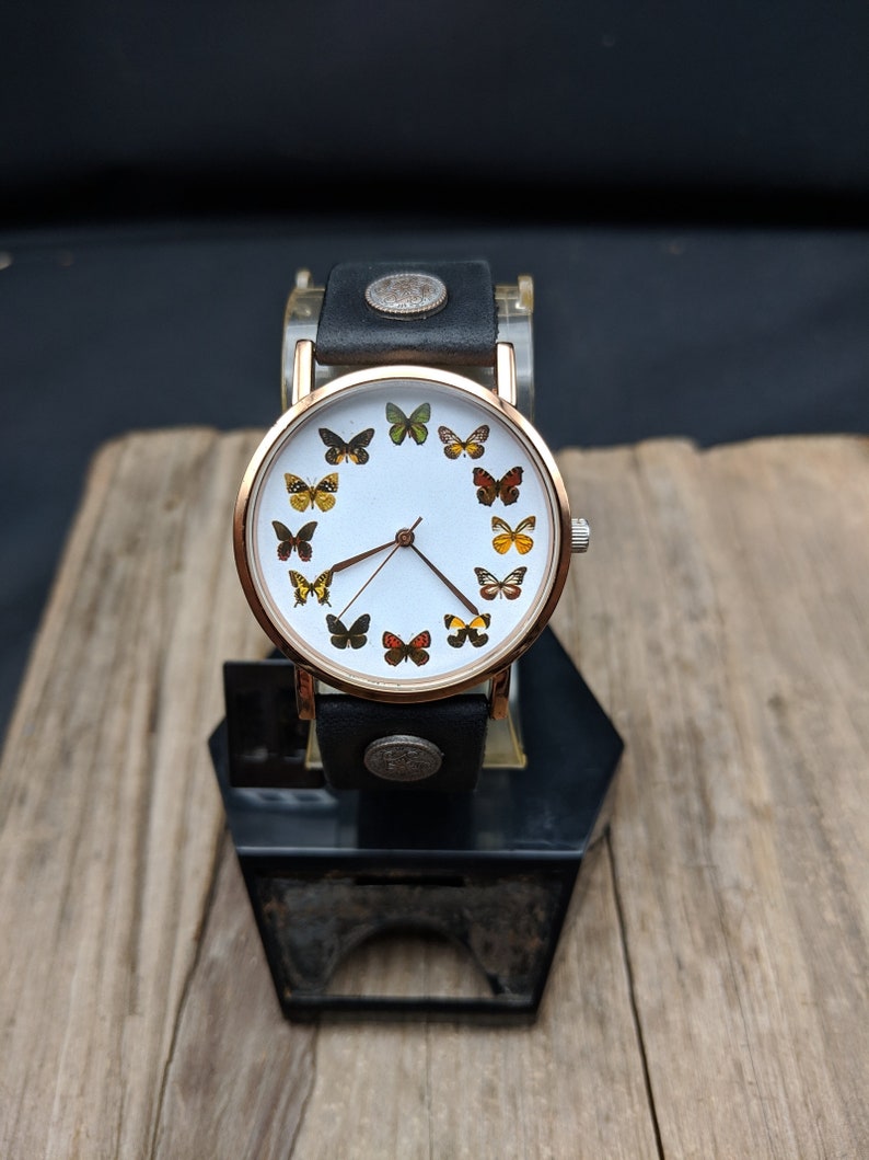 K&ouml;nnte beinhalten: Ein wei&szlig;es Zifferblatt mit einem schwarzen Lederarmband und goldenen Akzenten. Das Zifferblatt zeigt ein Design mit Schmetterlingen mit unterschiedlich gef&auml;rbten Fl&uuml;geln.