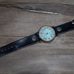 May include: A black leather wristwatch with a white face and roman numeral markings. The watch is on a wooden surface.