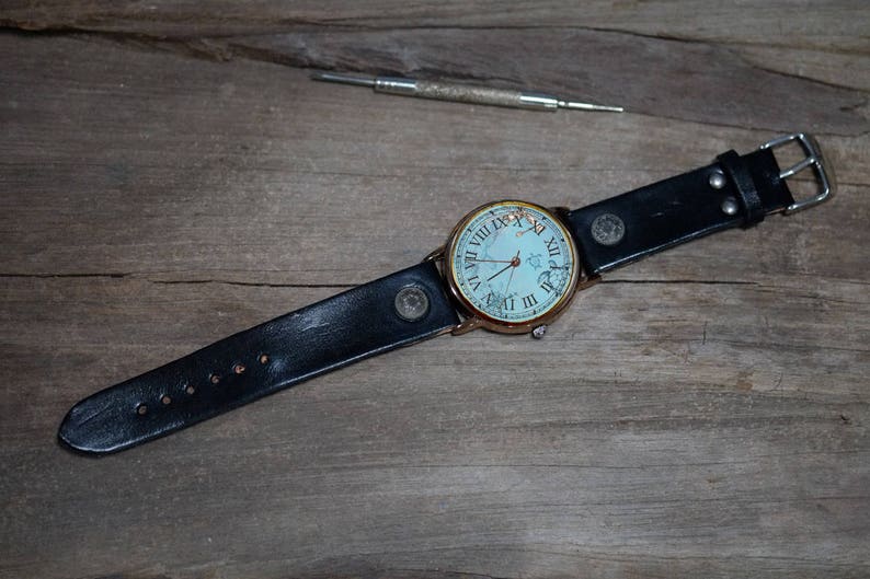May include: A black leather wristwatch with a white face and roman numeral markings. The watch is on a wooden surface.