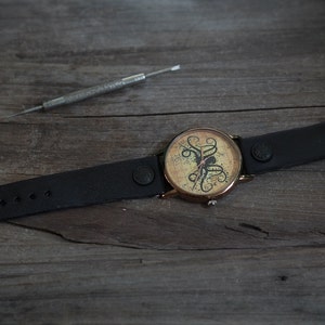 Pu&ograve; includere: Un orologio da polso in pelle nera con cassa dorata e quadrante bianco con un design di polpo nero. L'orologio ha una chiusura a fibbia.