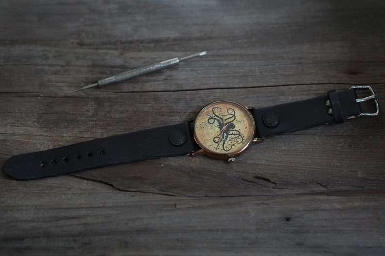 Pu&ograve; includere: Un orologio da polso in pelle nera con cassa dorata e quadrante bianco con un design di polpo nero. L'orologio ha una chiusura a fibbia.