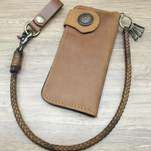 May include: A tan leather long wallet featuring a decorative antique brass concho snap closure, contrast stitching, and a braided leather wallet chain attached to a brass swivel clip and a leather belt loop strap with a brass snap button.