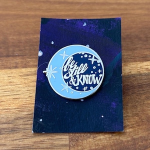 May include: A round enamel pin featuring a light blue crescent moon and a dark blue circle with white stars. The pin has the words "be still & KNOW" in white script. The pin is on a dark blue background.
