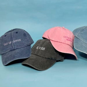 May include: Four baseball caps in blue, charcoal, pink, and light blue. The blue cap features the text OUT OF OFFICE, the charcoal cap says ILYSB, the pink cap reads LET'S GO GET MARGS, and the light blue cap displays the text Stay hydrated.