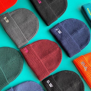 May include: A selection of ribbed knit beanies in colours like black, grey, navy, and burgundy. Each beanie has embroidered initials in contrasting colours, such as white, pink, and yellow. The beanies are arranged on a turquoise background.