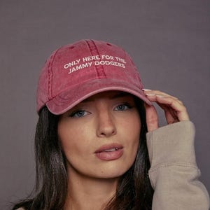 May include: A faded red baseball cap with the white text "ONLY HERE FOR THE JAMMY DODGERS". The cap is worn by a person wearing a beige jacket and a light brown shirt.