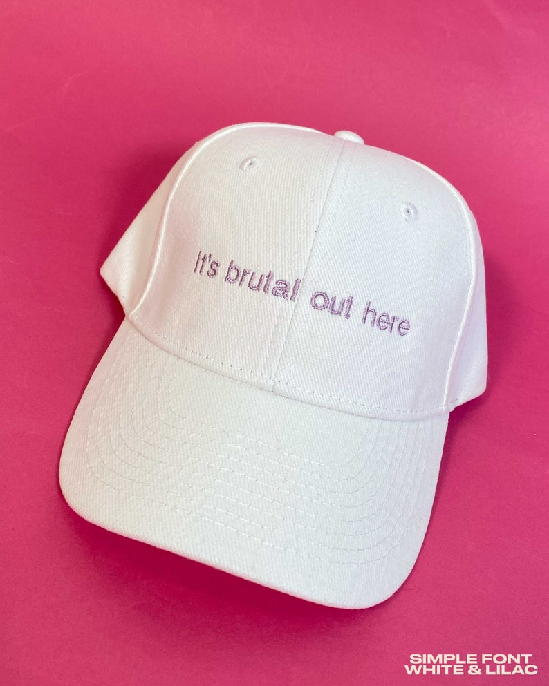 May include: A white baseball cap featuring the embroidered text "It's brutal out here" in a light purple thread. The cap has a structured crown, a curved brim with visible stitching, and a button at the top center of the fabric panels.