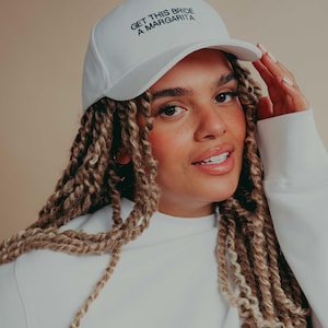 May include: A white baseball cap featuring the black embroidered text GET THIS BRIDE A MARGARITA on the front panel. The cap has a structured crown, a curved brim, and a matching white button on the top center of the headwear piece.