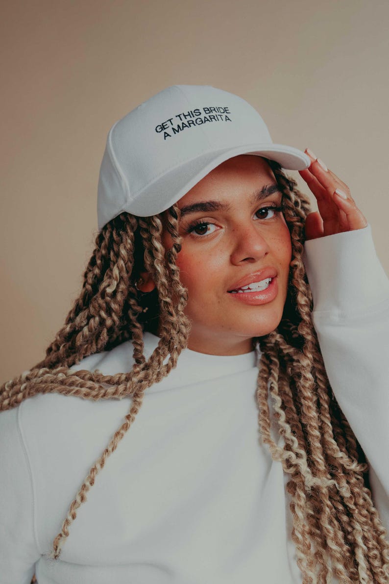 May include: A white baseball cap featuring the black embroidered text GET THIS BRIDE A MARGARITA on the front panel. The cap has a structured crown, a curved brim, and a matching white button on the top center of the headwear piece.