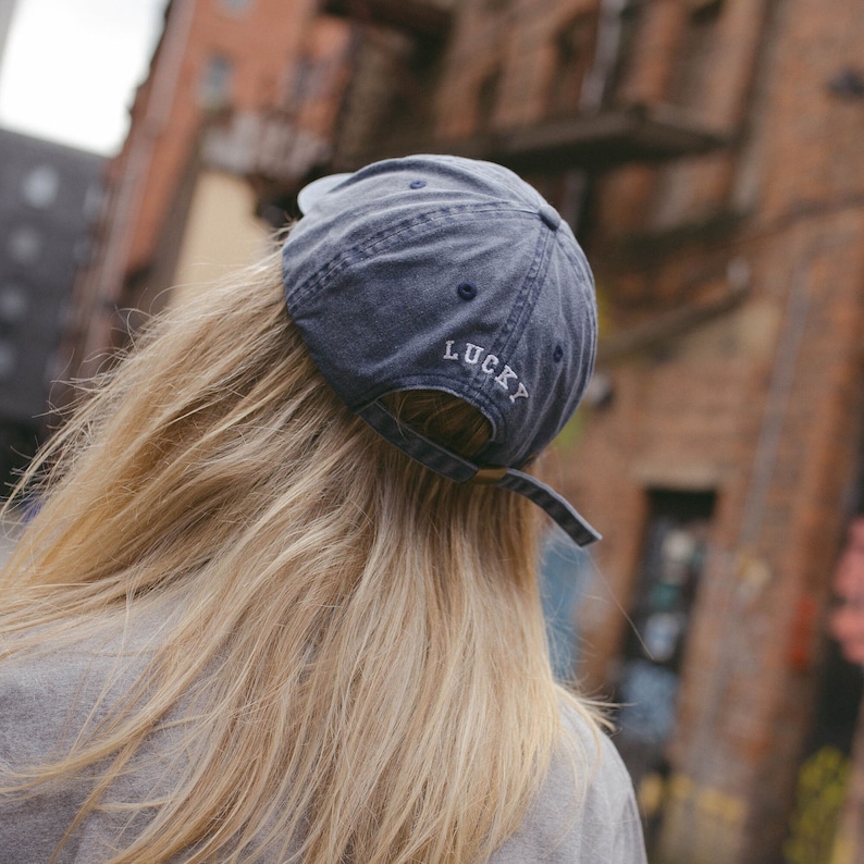 May include: A faded navy blue baseball cap with a six-panel construction and embroidered eyelets. The word LUCKY is stitched in white capital letters above the adjustable fabric strap closure, which features a metallic buckle at the back.