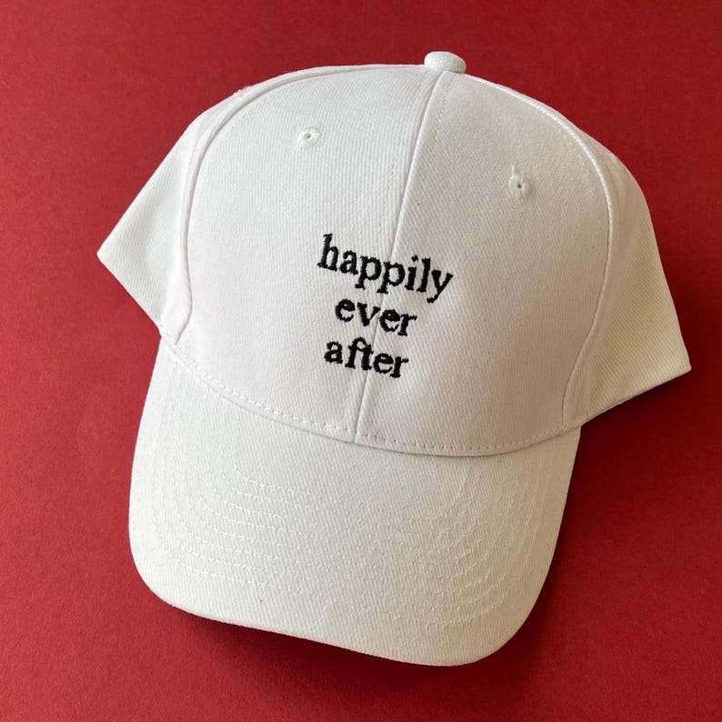 May include: A white baseball cap featuring the black embroidered text "happily ever after" centered on the front panel. The cap has a structured crown, a curved brim, and a button at the top, constructed from a solid white fabric material.