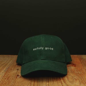 May include: A bottle green baseball cap featuring the text "awfully good" embroidered in white thread across the front. The cap has a structured crown and a curved brim, with the text "BOTTLE GREEN CAP WHITE THREAD SIMPLE FONT" visible.