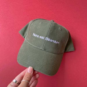 May include: An olive green baseball cap featuring the white embroidered text Nunc est Bibendum on the front panel. The cap has a structured crown, a curved brim, and stitched eyelets for ventilation, constructed from a durable fabric material.