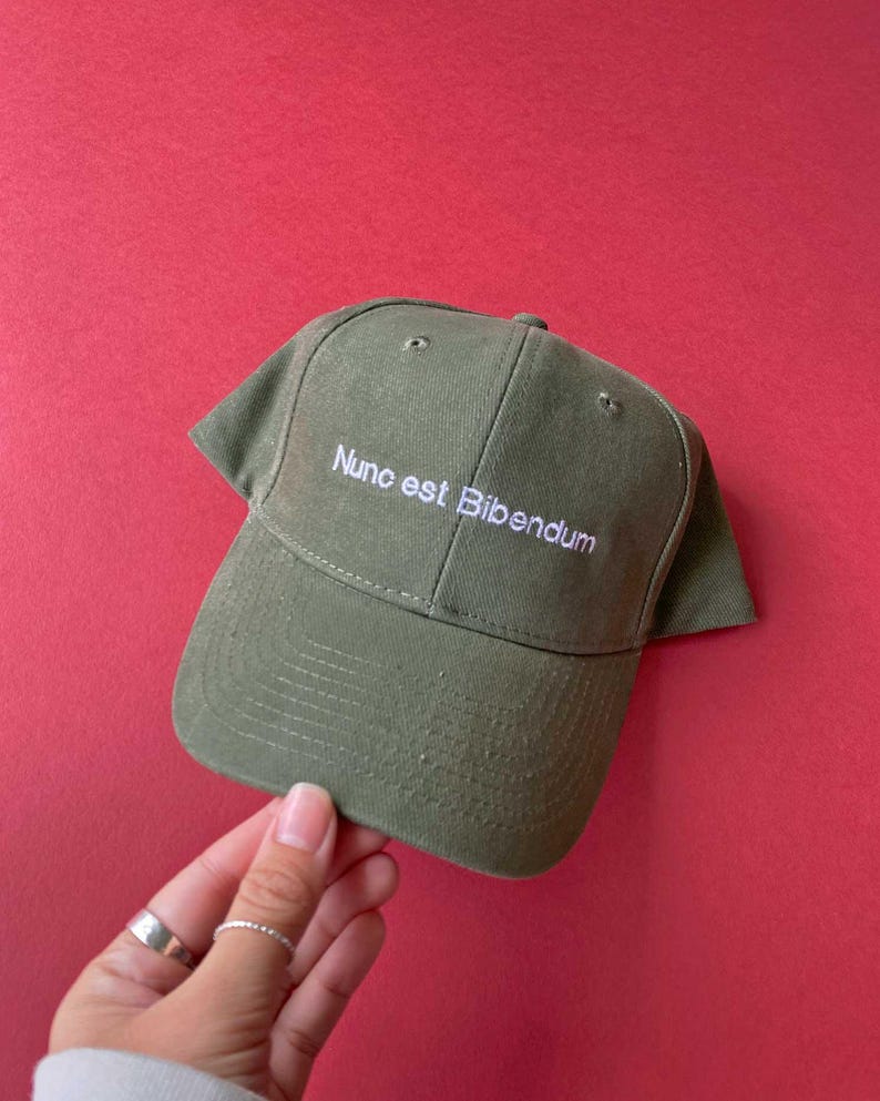 May include: An olive green baseball cap featuring the white embroidered text Nunc est Bibendum on the front panel. The cap has a structured crown, a curved brim, and stitched eyelets for ventilation, constructed from a durable fabric material.