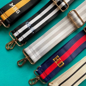 May include: Five different striped fabric straps with gold clasps. The straps have different colours and patterns, including black and white, grey and white, navy blue and red, and yellow and black. The straps are personalised with embroidered text, including "MRS", "03.11", and "HD".