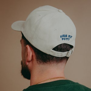 May include: A beige baseball cap with a blue embroidered message that reads "Kiss My Putt".
