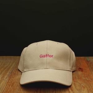 May include: A cream-colored baseball cap featuring the word Gaffer embroidered in hot pink thread using a simple font. The cap has a structured crown and a curved brim, with visible stitching details across the panels and the front embroidery.
