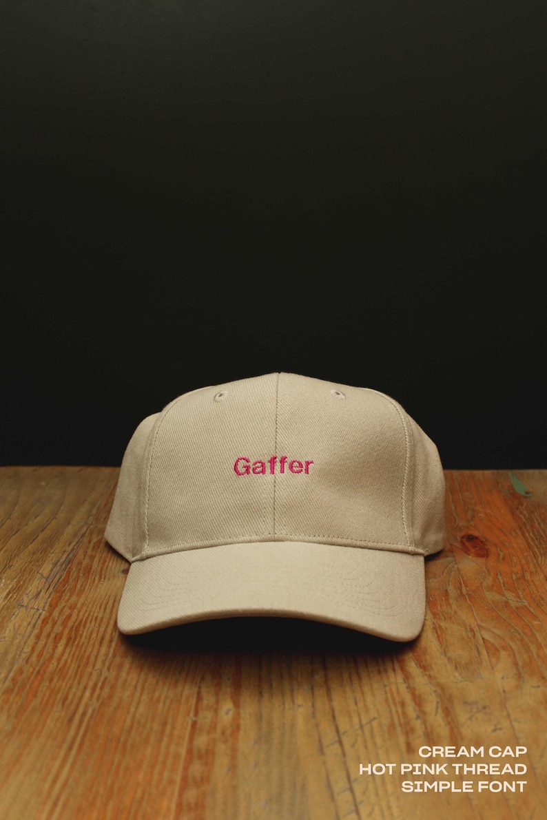 May include: A cream-colored baseball cap featuring the word Gaffer embroidered in hot pink thread using a simple font. The cap has a structured crown and a curved brim, with visible stitching details across the panels and the front embroidery.