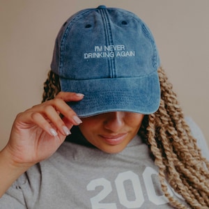 May include: A blue denim baseball cap with a distressed, washed-out finish. The front of the cap features the embroidered white text "I'M NEVER DRINKING AGAIN" in a clean, sans-serif font. The cap has a curved brim and stitched eyelets.