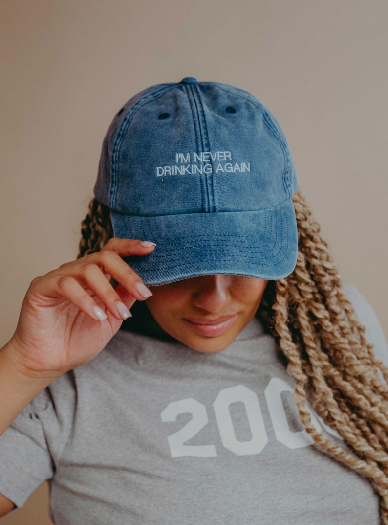 May include: A blue denim baseball cap with a distressed, washed-out finish. The front of the cap features the embroidered white text "I'M NEVER DRINKING AGAIN" in a clean, sans-serif font. The cap has a curved brim and stitched eyelets.