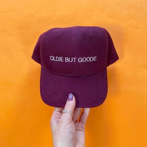 May include: A deep burgundy baseball cap featuring the white embroidered text "OLDIE BUT GOODIE" centered on the front panel. The cap has a structured crown, a curved brim, and small ventilation eyelets on the top panels of the fabric.