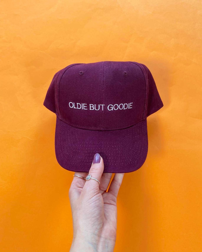 May include: A deep burgundy baseball cap featuring the white embroidered text "OLDIE BUT GOODIE" centered on the front panel. The cap has a structured crown, a curved brim, and small ventilation eyelets on the top panels of the fabric.