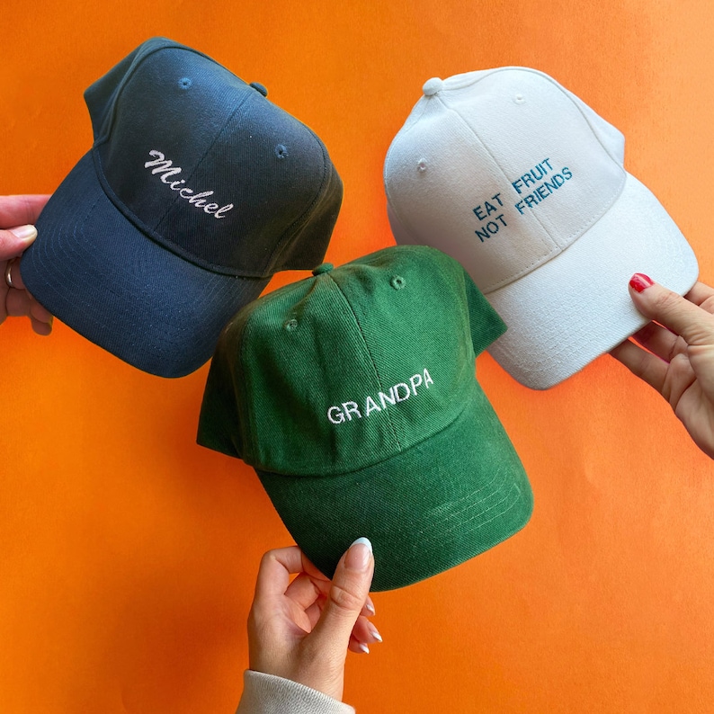 May include: Three baseball caps in navy blue, forest green, and white. The navy cap features the embroidered name Michel. The green cap displays the word GRANDPA in white embroidery. The white cap features the text EAT FRUIT NOT FRIENDS.