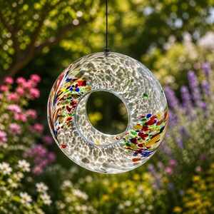 May include: A circular glass bird feeder featuring a clear, textured surface with embedded multi-colored flecks in shades of red, blue, yellow, and green. The hollow center allows for bird seed, and it hangs from a thin black cord.