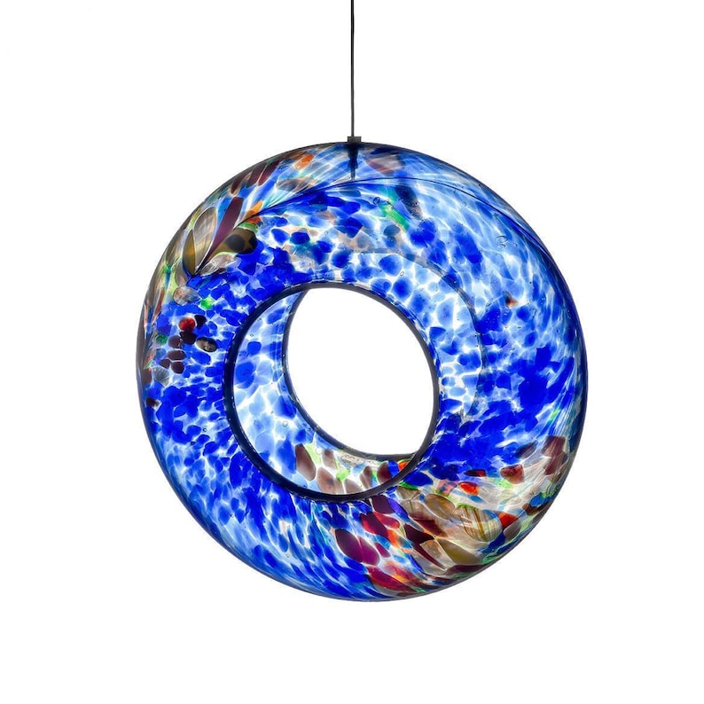 May include: A circular, hollow glass ornament featuring a vibrant blue speckled pattern with accents of white, red, and brown. The piece is suspended by a thin black cord, showcasing a smooth, polished surface and a central open void.