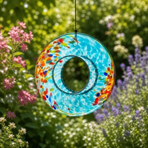May include: A circular glass bird feeder featuring a vibrant turquoise blue base with a speckled pattern of red, orange, yellow, and white glass fragments. The feeder has a central circular opening and is suspended by a thin black cord.