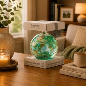 May include: A spherical glass ornament with swirling green, blue, and gold patterns inside a clear box. The packaging is labelled Angel Orb Archangel Raphael and The Sienna Collection. The box is placed on a wooden surface next to a candle and books.