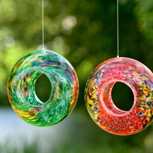 May include: Two circular glass ornaments hanging from thin strings. The left ornament features a swirling pattern of green, yellow, and blue glass. The right ornament displays a speckled design with vibrant red, orange, yellow, and blue glass colors.