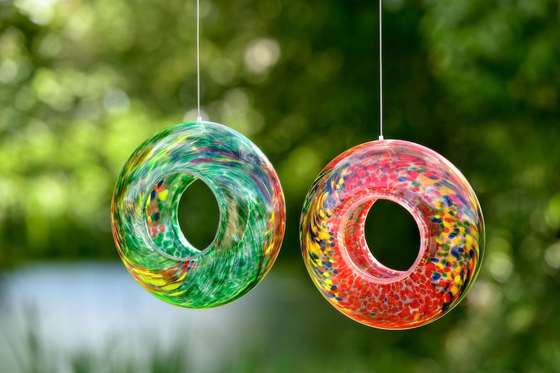 May include: Two circular glass ornaments hanging from thin strings. The left ornament features a swirling pattern of green, yellow, and blue glass. The right ornament displays a speckled design with vibrant red, orange, yellow, and blue glass colors.