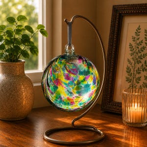 May include: A spherical glass ornament featuring a vibrant, multi-coloured pattern of pink, yellow, green and blue swirls. The ornament hangs from a grey ribbon attached to a curved, bronze-toned metal stand with a circular base.