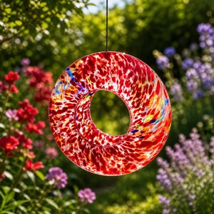 May include: A circular glass suncatcher featuring a vibrant speckled pattern of red, orange, yellow, and blue colors. The piece has a hollow center and is suspended by a thin black cord, allowing light to pass through the translucent glass surface.