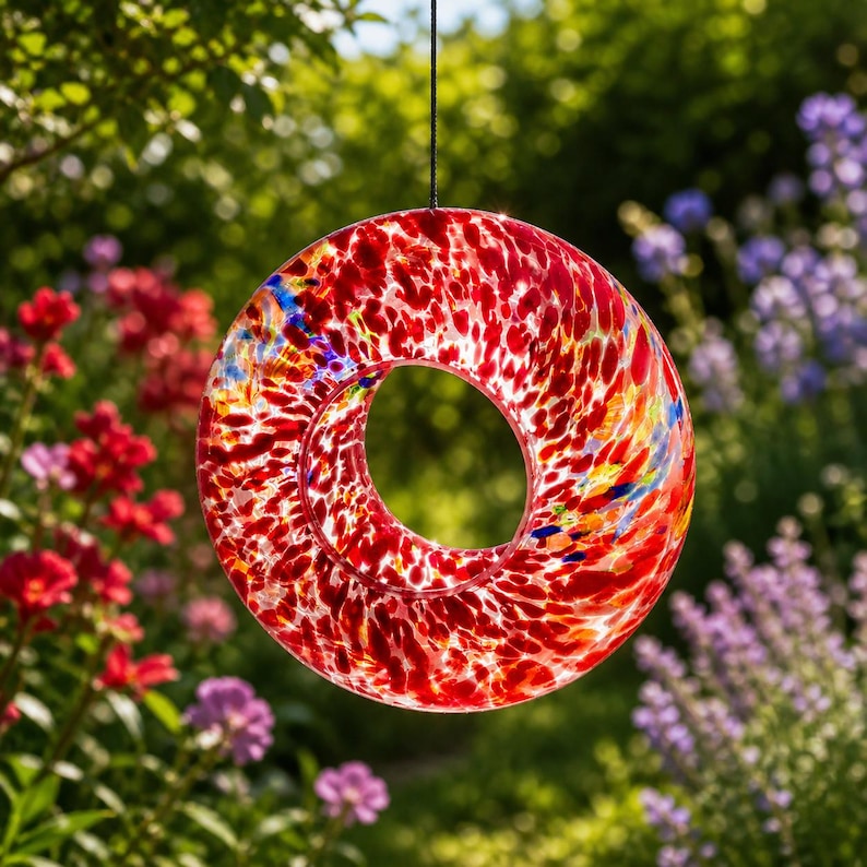 May include: A circular glass suncatcher featuring a vibrant speckled pattern of red, orange, yellow, and blue colors. The piece has a hollow center and is suspended by a thin black cord, allowing light to pass through the translucent glass surface.