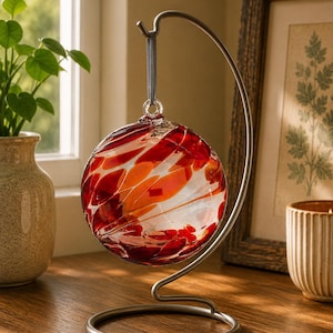 May include: A spherical glass ornament featuring swirling patterns of deep red and orange hues suspended from a curved, brushed metal stand. The ornament hangs by a thin grey ribbon attached to a small glass loop at the top of the sphere.